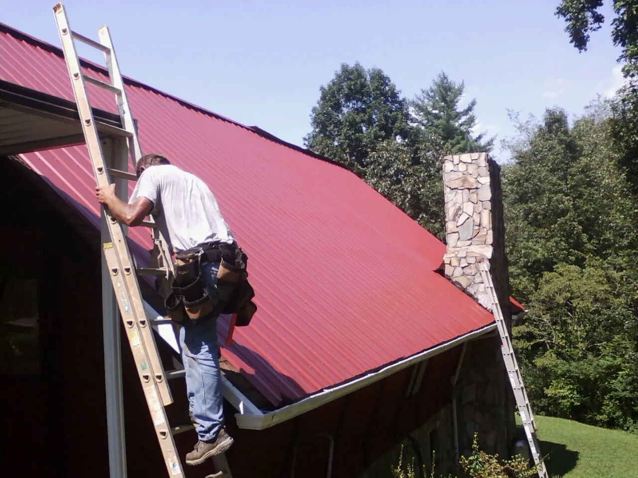 Roofer on a ladder beside a red metal roof and stone chimney — Soffit & Fascia Repair by Roofing Brothers in Chelan Falls